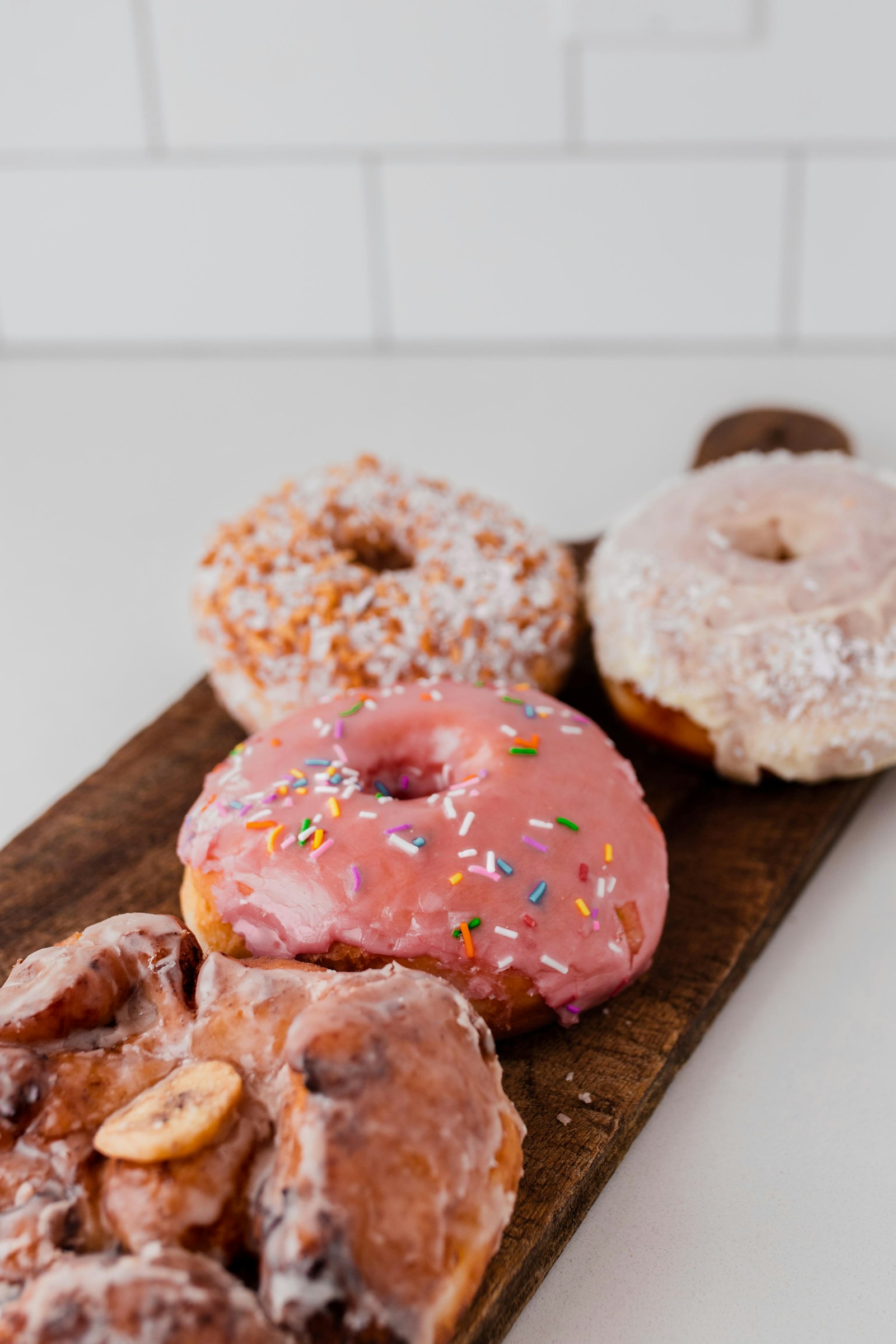 A pink-glazed sprinkle donut on a wooden board with coconut donuts and a glazed twist